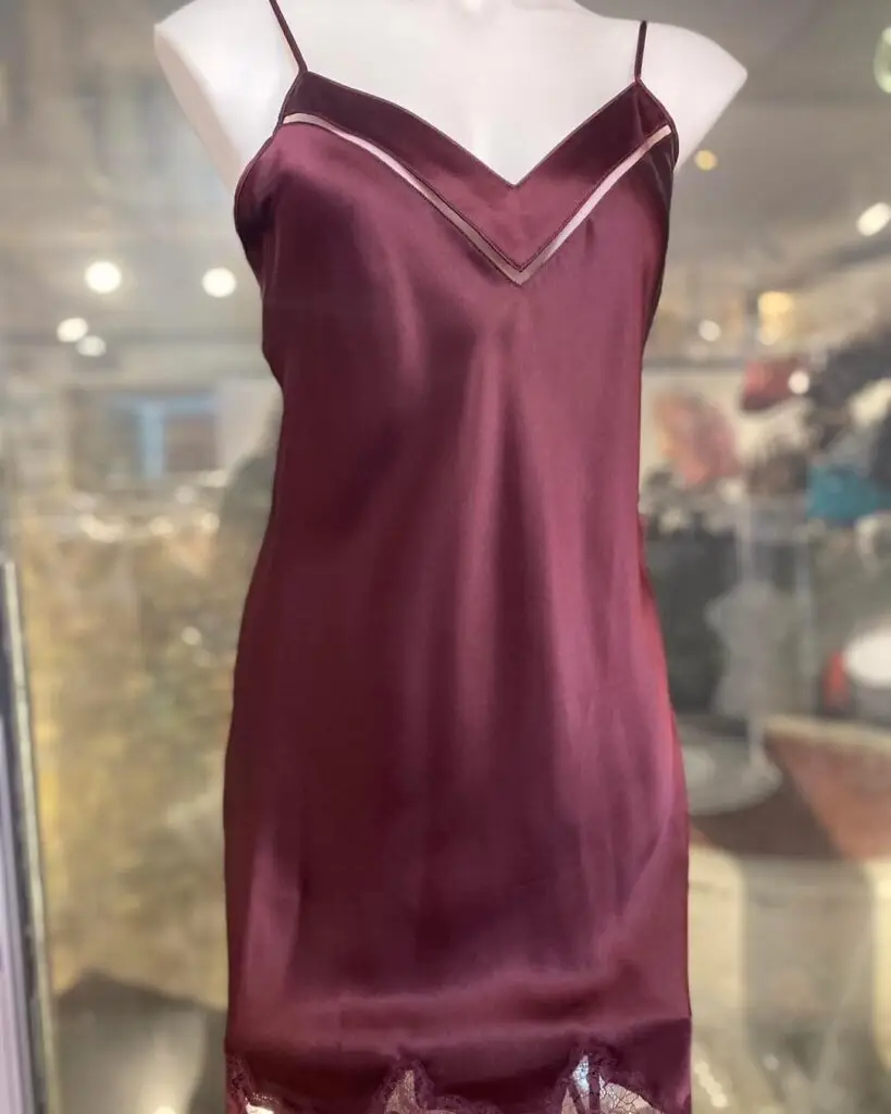 A rich burgundy satin chemise featuring a V-neck design with sheer accents and delicate lace trim at the hem, displayed on a mannequin in a boutique setting.